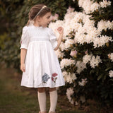 Little girl wearing a white embroidered dress standing beside blooming flowers
