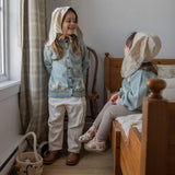 Two children in vintage-style clothing with headscarves in a room with wooden furniture and a horse painting on the wall.