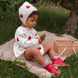 A toddler sits on a beige blanket outdoors in a grassy garden, wearing a white knit hooded romper with red apple prints and bright red shoes. A wicker basket is nearby, with trees and greenery in the background.