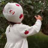 A child seen from behind wearing a cream-colored knitted bonnet and matching long-sleeve dress, both decorated with red embroidered apple motifs, standing outdoors in front of green foliage.