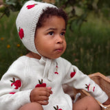A baby wearing a white knit bonnet and matching long-sleeve romper, both decorated with red apple motifs, sitting outdoors in a wicker basket.