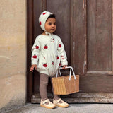 A toddler stands in front of a rustic dark wooden door wearing a cream-colored strawberry-print romper, matching bonnet, brown leggings, and beige shoes while holding a small woven basket.
