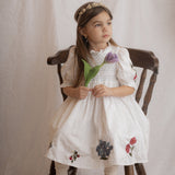 Young girl in a white dress with floral embroidery sitting on a wooden chair.