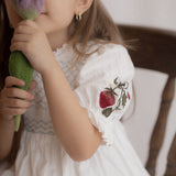 Young girl in a white dress with floral embroidery holding a green plush toy.