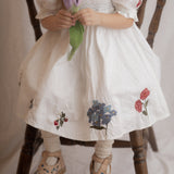 Child wearing a white dress with floral embroidery, sitting on a wooden chair.