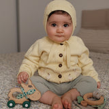A baby sitting on a speckled carpet wearing a pale yellow knit bonnet and matching cardigan with gray pants, holding a wooden toy car.