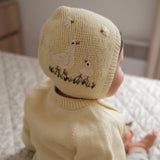 A baby wearing a cream-colored knit hat with embroidered swans and a cream knit sweater, sitting on a patterned carpet.