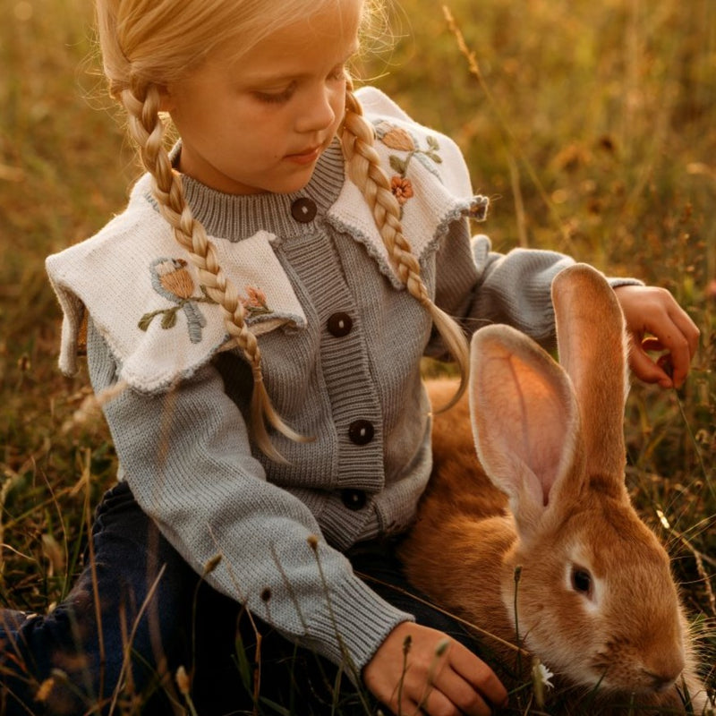 A little girl playing with an animal in a meadow, surrounded by greenery and nature, wearing the Little Birdie cardigan.







