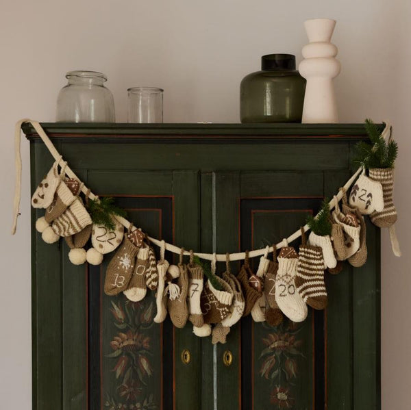 Cream wool Advent Calendar, featuring 24 handcrafted mittens, hats, and socks, hung on a decorated wardrobe with festive ornaments.