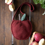 A crocheted apple-shaped shoulder bag in deep burgundy red, featuring a small light green leaf accent on the stem, displayed on a dark wooden background with decorative apples and green leaves.