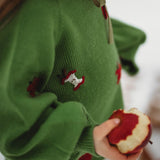 A close-up lifestyle shot of a child wearing a green knit sweater with red embroidered apple motifs, holding a bitten red apple in a snowy setting.