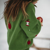 A child wearing a green knit sweater with red apple patches and a snowman applique, standing outdoors in a snowy setting.