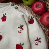 An off-white knit baby sweater with embroidered red apples and apple core motifs, styled with fresh red and green apples on a woven mat.