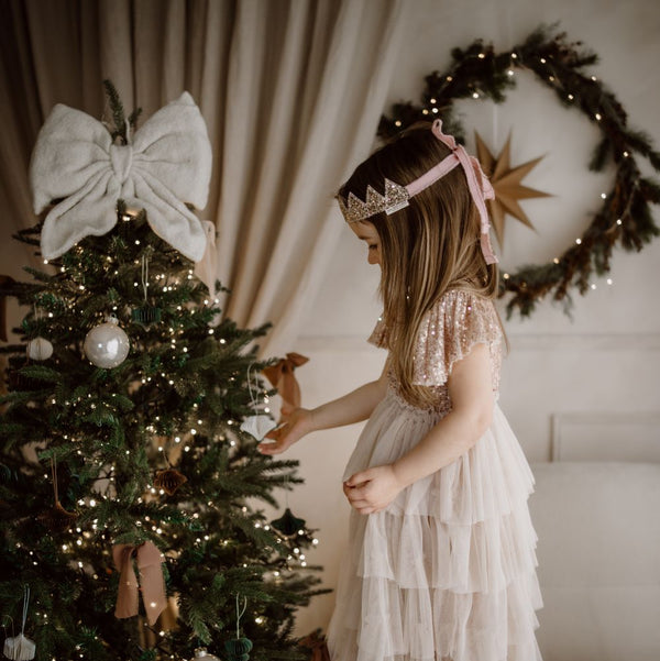 Young girl in a white dress standing next to a decorated Christmas tree with a wreath in the background.