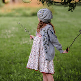 Young girl in a floral dress and blue cardigan standing in a grassy field holding sticks.
