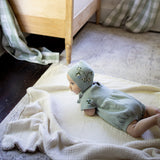 Baby in a green outfit and hat lying on a bed in a softly lit room.