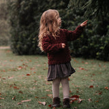 Child in a red sweater and skirt standing in a grassy area with trees in the background