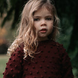 Young girl wearing a dark red textured sweater outdoors with blurred greenery in the background