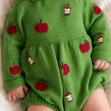 A close-up of a baby wearing a bright green knit romper decorated with red apple motifs and small white-and-red apple slice patches, lying on a soft cream fabric.