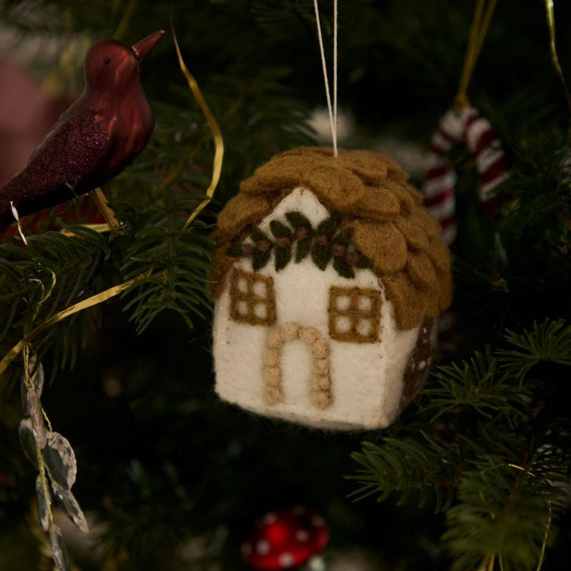 Handcrafted white gingerbread house ornament hanging on a Christmas tree, surrounded by other festive decorations. The ornament features intricate details and is made from 100% wool, contributing to the holiday cheer with its traditional and whimsical design.