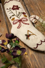 Knitted stocking with floral and bow design on a wooden surface with flowers.