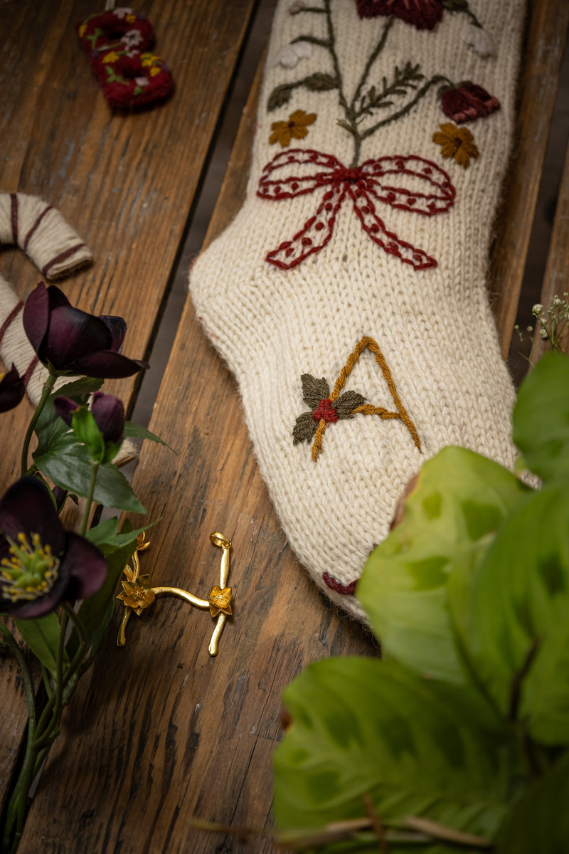 Knitted sock with floral embroidery and a gold 'A' on a wooden surface with flowers.