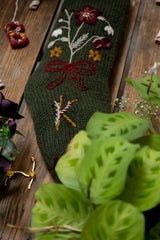 Green knitted stocking with floral and bow design on a wooden surface with plants.
