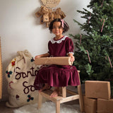 a girl sitting next to a christmas tree with a santa sack next to it