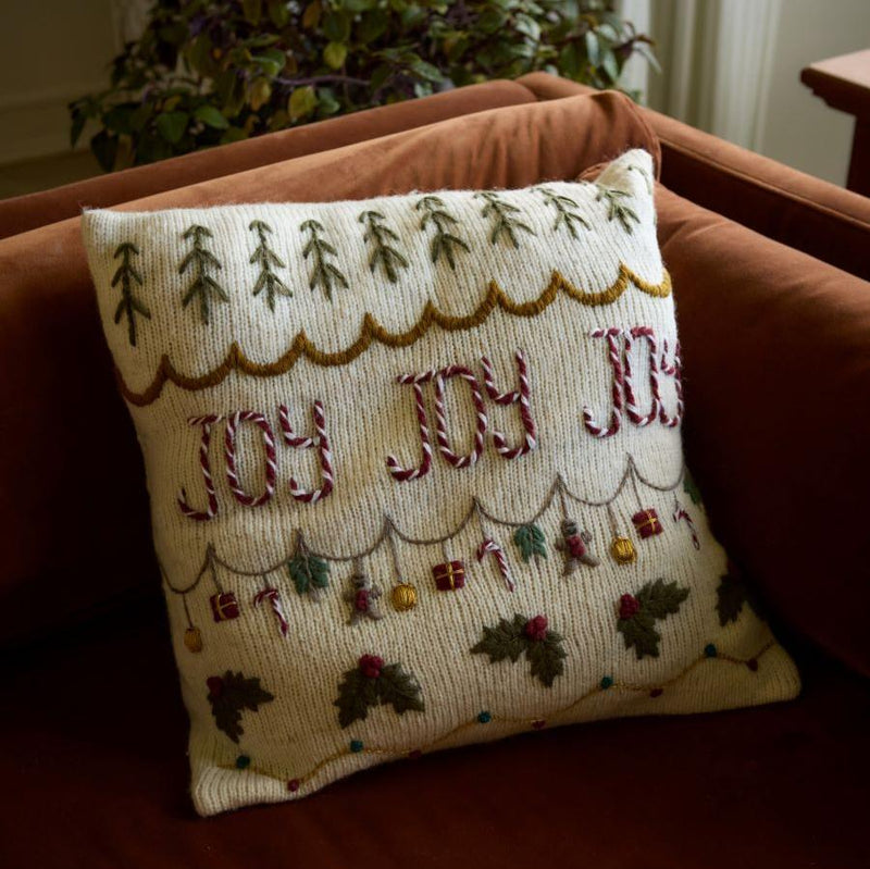 Joy Pillow Cover in Cream White displayed on a sofa, adding a festive and cozy touch with its hand-knitted texture and embroidered JOY design.