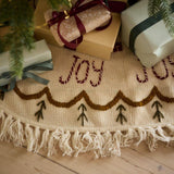 Joy Heirloom Tree Rug displayed around a Christmas tree, adding a festive and elegant touch with its hand-embroidered design and luxurious knitted texture.