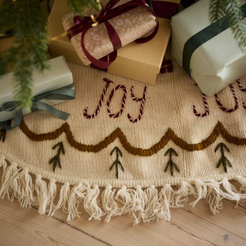 Joy Heirloom Tree Rug displayed around a Christmas tree, adding a festive and elegant touch with its hand-embroidered design and luxurious knitted texture.