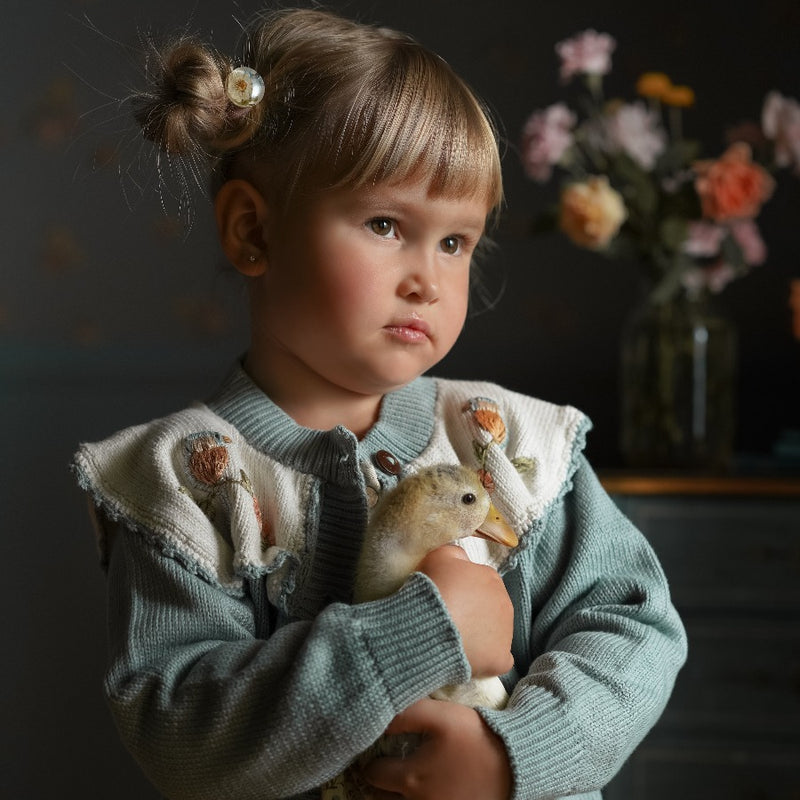 Girl wearing the Little Birdie cardigan, holding an animal, with flowers softly blurred in the background.







