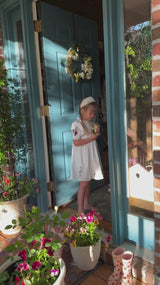 
Child wearing a white embroidered dress standing in a doorway surrounded by flowers