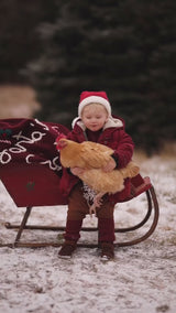 A child feeding chickens while a red wool Santa sack with "Santa's Sack" embroidered is placed on a sled nearby.