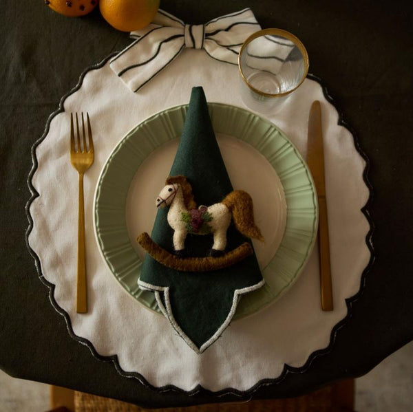 Hand-felted rocking horse ornament displayed on a table, placed on a plate, and positioned on top of a napkin. The ornament adds a whimsical touch to the holiday table setting.