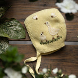 Yellow knitted baby bonnet with bird design on a wooden surface with flowers and leaves.