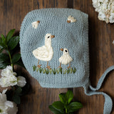 Knitted baby hat with duck design on a wooden surface surrounded by flowers