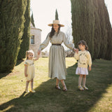 Woman in a long dress and hat walking with two children in a sunlit outdoor setting with tall trees.