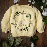 Yellow sweater with floral and bird design on a wooden surface with flowers and leaves.