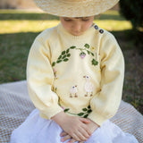 Child wearing a yellow sweater with bird design sitting on a blanket outdoors.