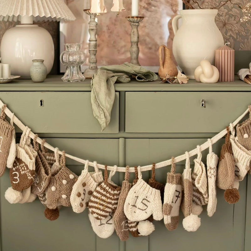 Cream wool Advent Calendar hanging on a sideboard, displaying the handcrafted mittens, hats, and socks with embroidered numbers.