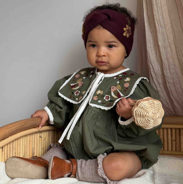 baby sitting in a sewn green romper with embroidered collar and grape hairband