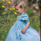 Young girl in a blue dress standing on a wooden path with flowers and greenery around.