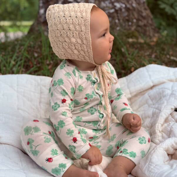 Baby in a knitted bonnet and pajamas with a clover pattern sitting on a blanket outdoors.