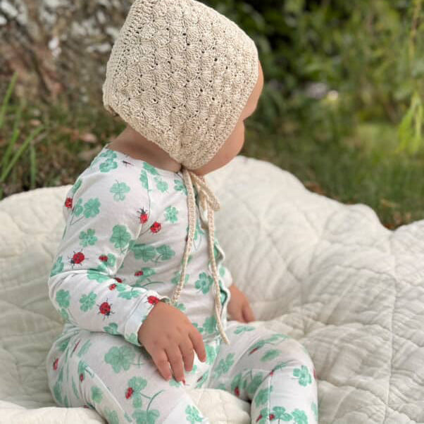Baby in a clover outfit and knitted hat sitting on a blanket outdoors.