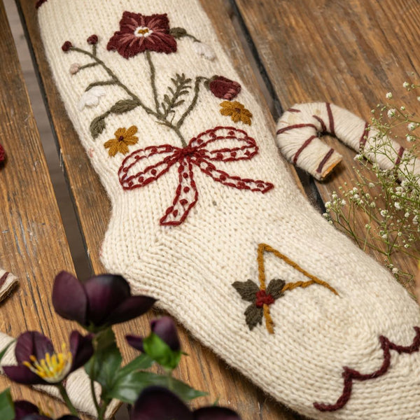 Knitted stocking with floral and bow design on a wooden surface with flowers.