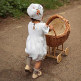 Child in a white dress and bonnet pushing a wooden pram outdoors.