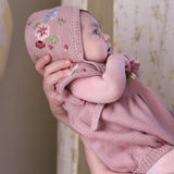 Baby wearing a pink knitted outfit with floral embroidery, held by an adult against a neutral background.
