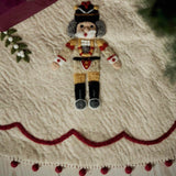 A felt Christmas nutcracker decoration placed on a cream-colored felt tree skirt with red scalloped trim and pom-poms, surrounded by holiday greenery and a wrapped gift.