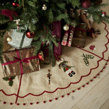 A festive Christmas tree skirt with embroidered nutcracker and holly designs, spread out on a light wood floor beneath a decorated Christmas tree with wrapped presents.